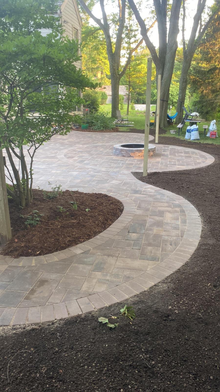 Winding brick path to outdoor patio with a circular fireplace in the back yard surrounded by forest.