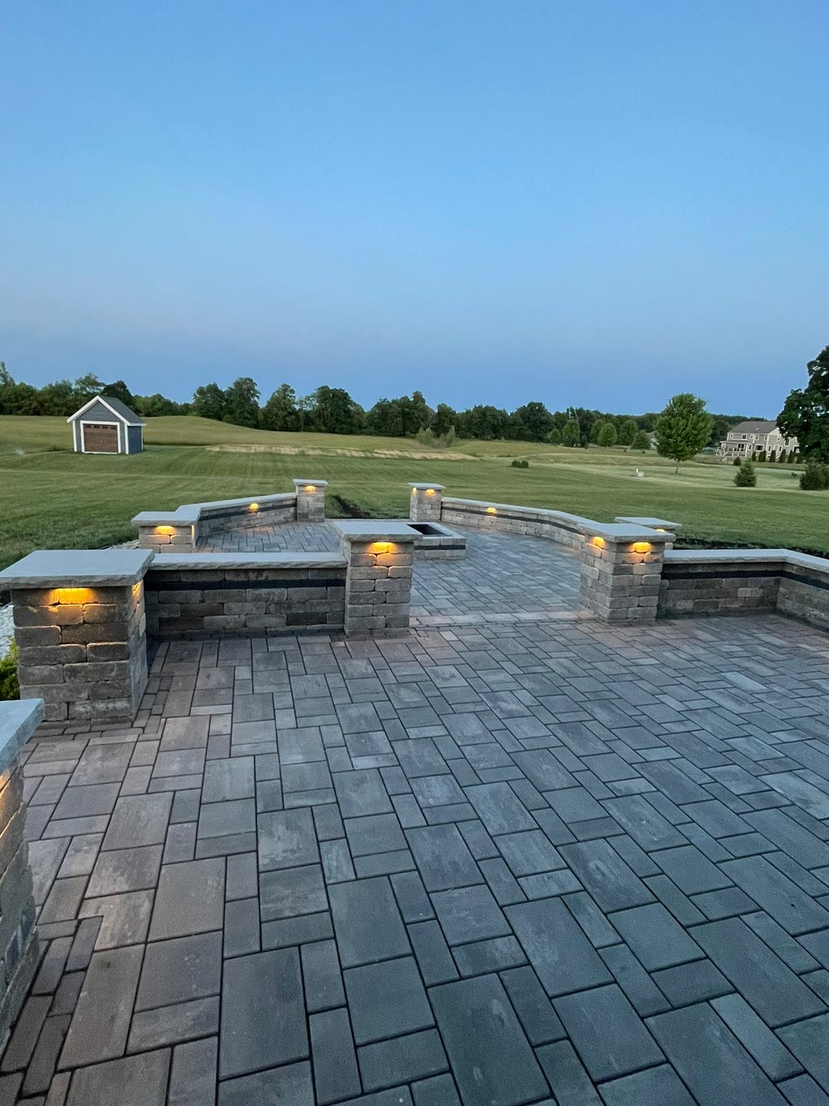 Multi-level natural stone patio with fieldstone retaining walls and pillars with lighting, centerpiece of a square fire pit. 