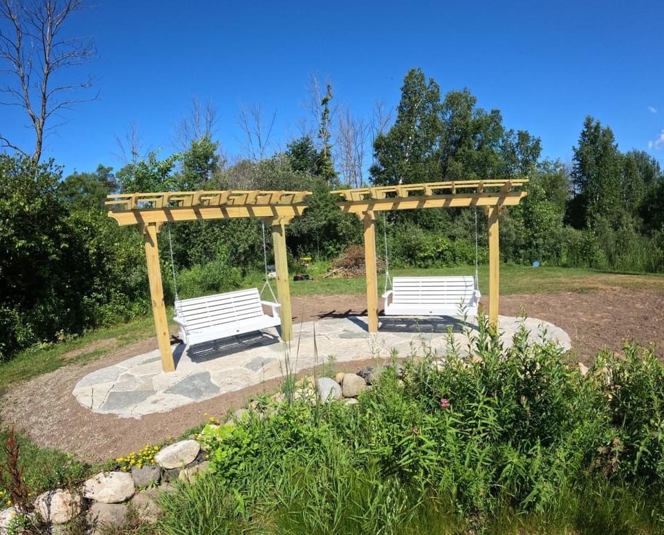 Two pergola swing benches on a flagstone base. Perfect for gazing at a pond, in front of a stone retaining wall, against a forest backdrop.